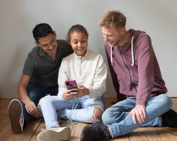 Drei Jugendliche sitzen nebeneinander auf einem Zimmerboden und schauen auf ein Smartphone, dass die Person in der Mitte hält